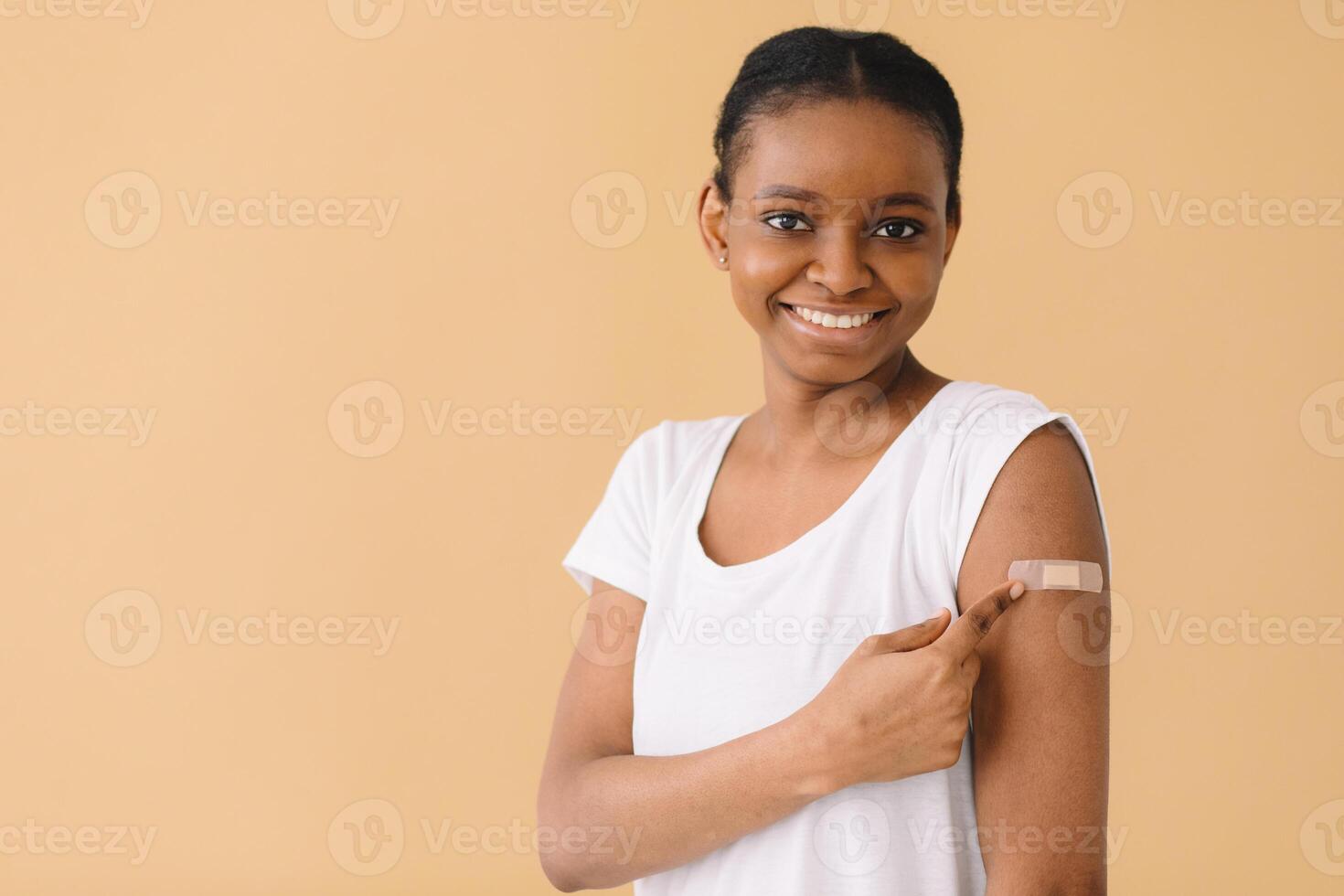 Young African American woman is happy about the first vaccination, wears band aid, raises sleeve and tells friends about the injection. Isolated over beige background. photo