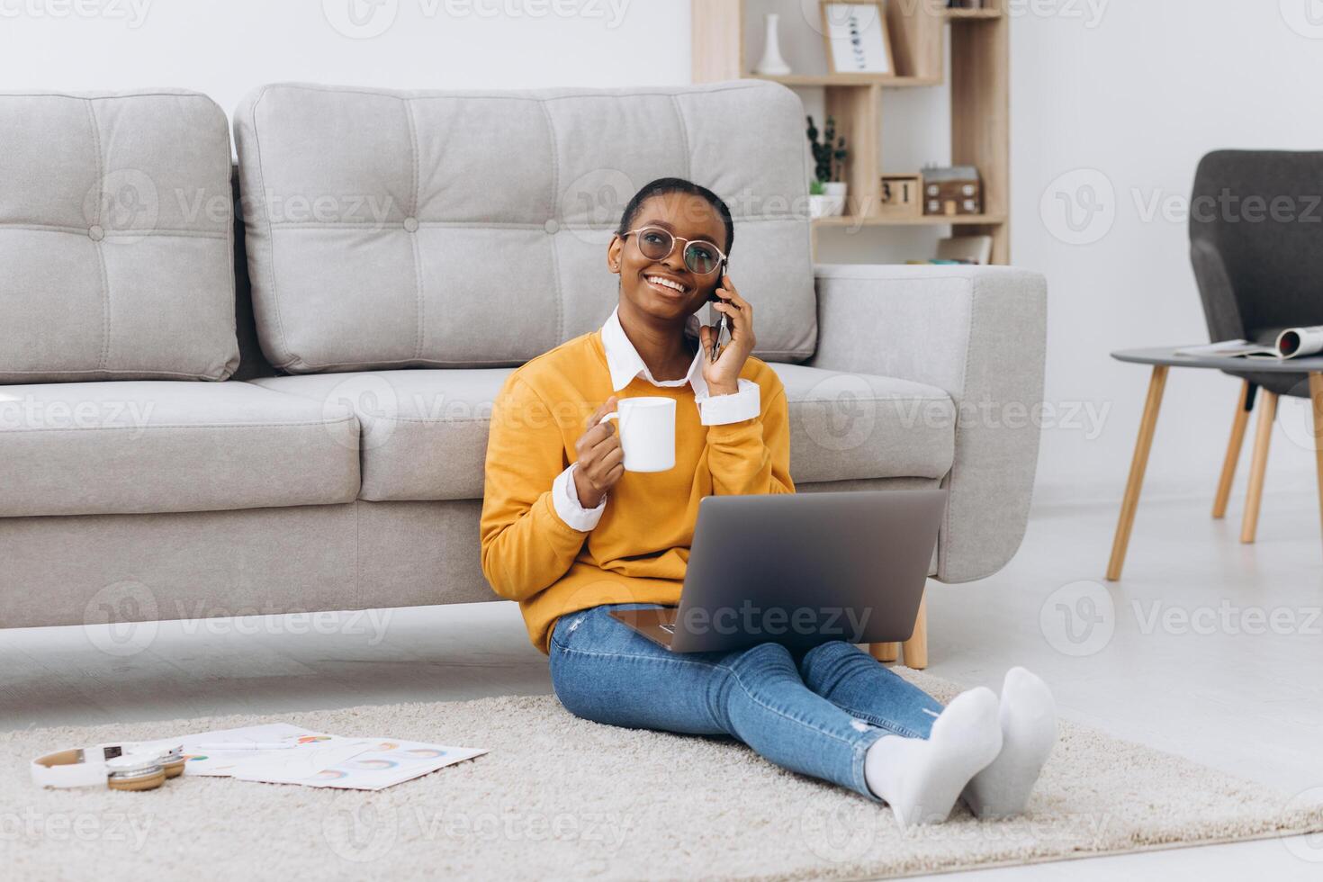 Beautiful young black university student sitting on the floor drinking coffee, talking on the phone and doing a project on a laptop photo