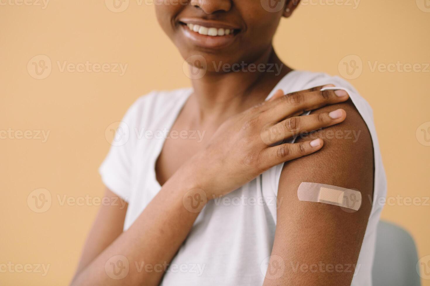 Young African American woman is happy about the first vaccination, wears band aid, raises sleeve and tells friends about the injection. Isolated over beige background. photo