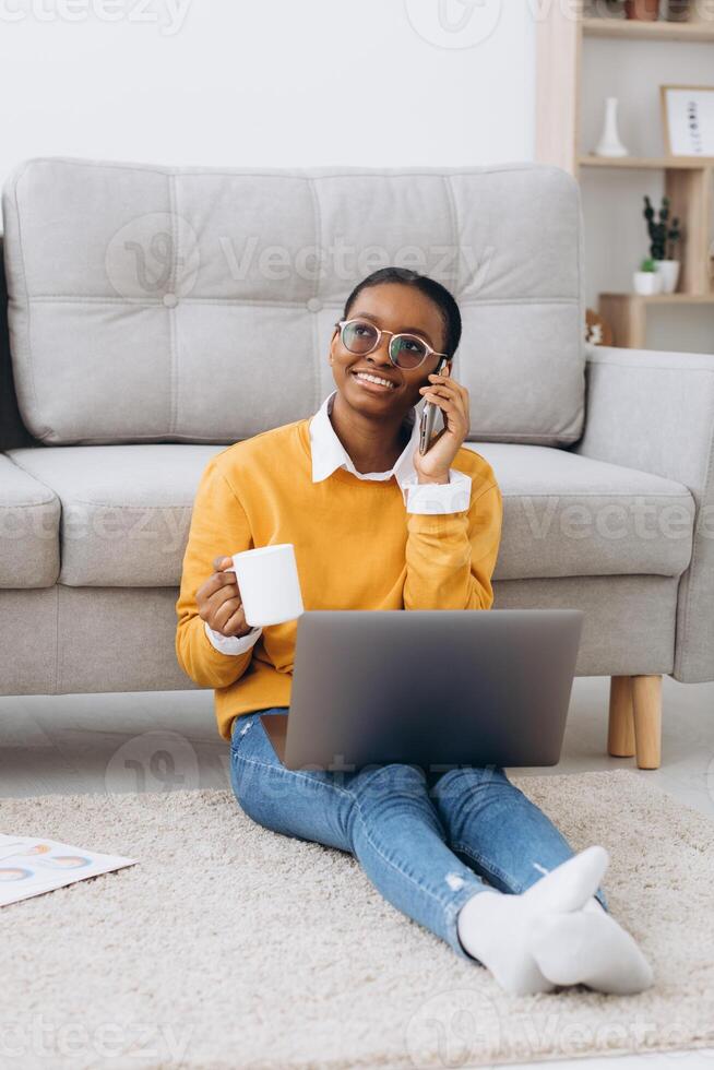 Beautiful young black university student sitting on the floor drinking coffee, talking on the phone and doing a project on a laptop photo