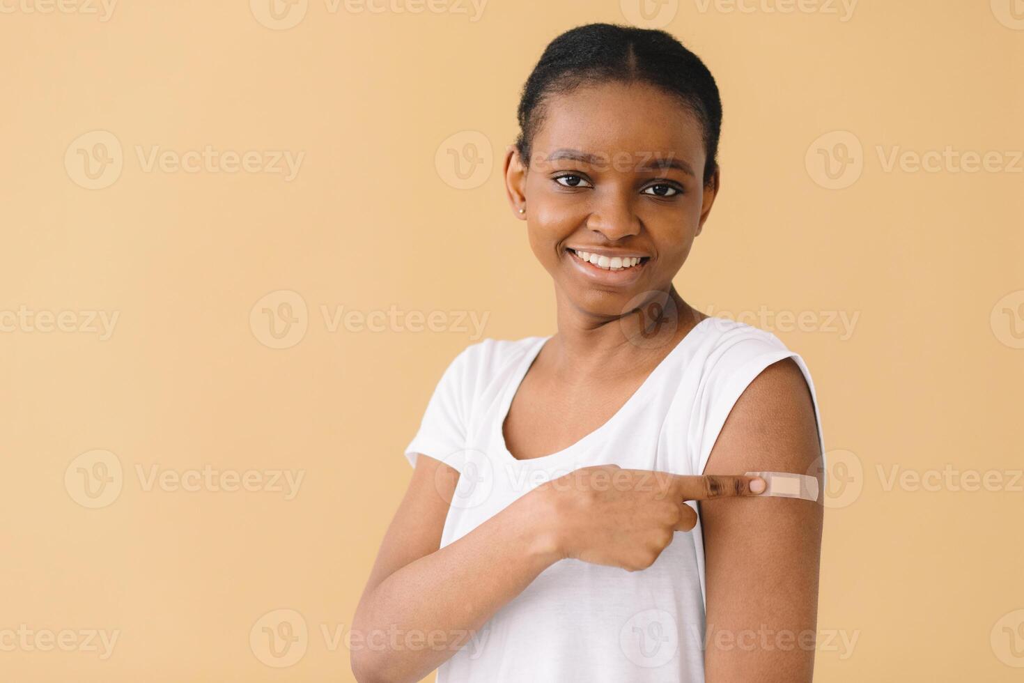 Young African American woman is happy about the first vaccination, wears band aid, raises sleeve and tells friends about the injection. Isolated over beige background. photo