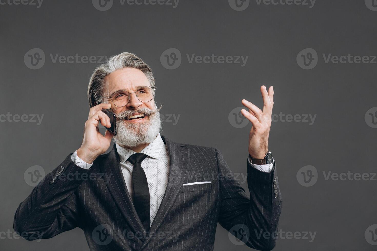 Business and fashionable bearded mature man in a gray suit smiling, talking by phone and showing copy space on the grey background. photo