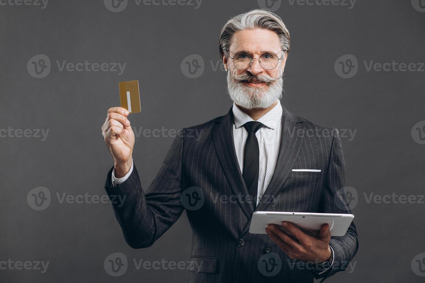 Business and fashionable bearded mature man in a gray suit making shopping in tablet with his gold credit card on the grey background. photo