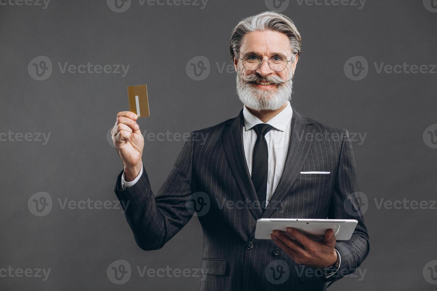 Business and fashionable bearded mature man in a gray suit making shopping in tablet with his gold credit card on the grey background. photo