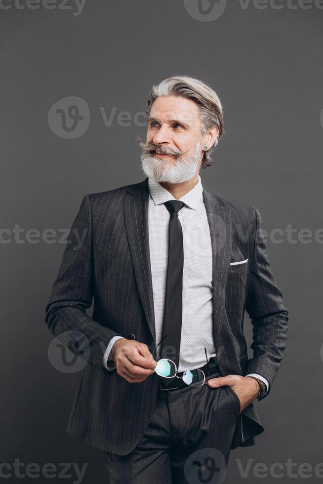 Business and fashionable bearded mature man in a gray suit holding his glasses and smiling on a grey background. photo