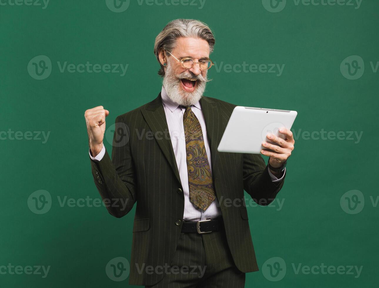 Bearded middle-aged man in khaki suit screaming loudly and looking at tablet while holding fist of happiness against green background photo