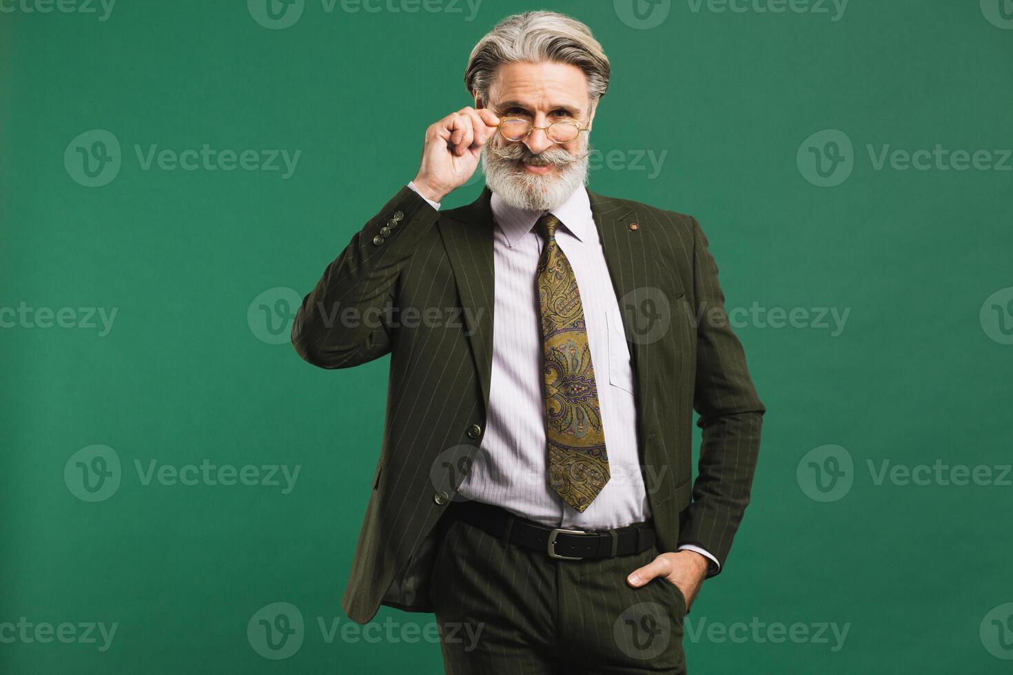 Stylish middle-aged bearded man in khaki suit holding glasses over green background photo