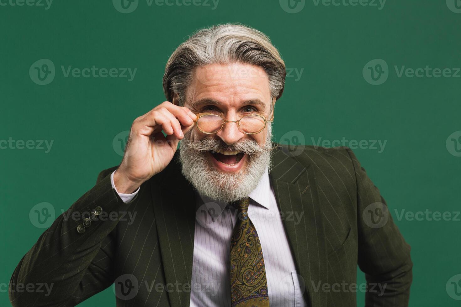 Bearded middle age teacher in khaki suit holds glasses with his hands and blinks his eyes with fun on green background photo