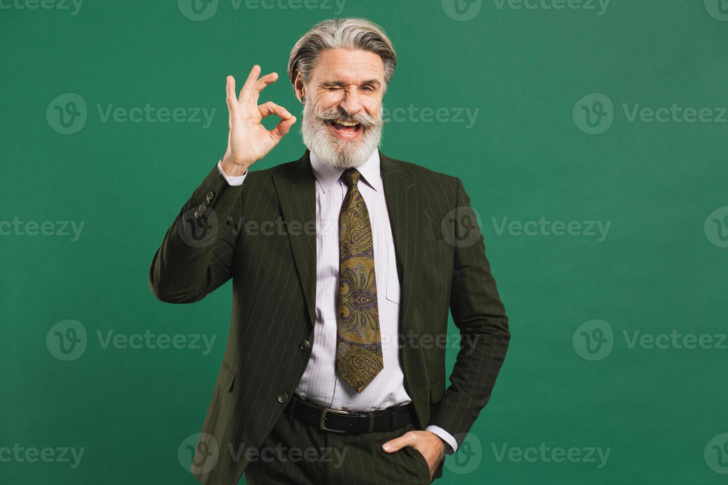 Bearded middle-aged man in khaki suit shows okay on green background with copy space. photo