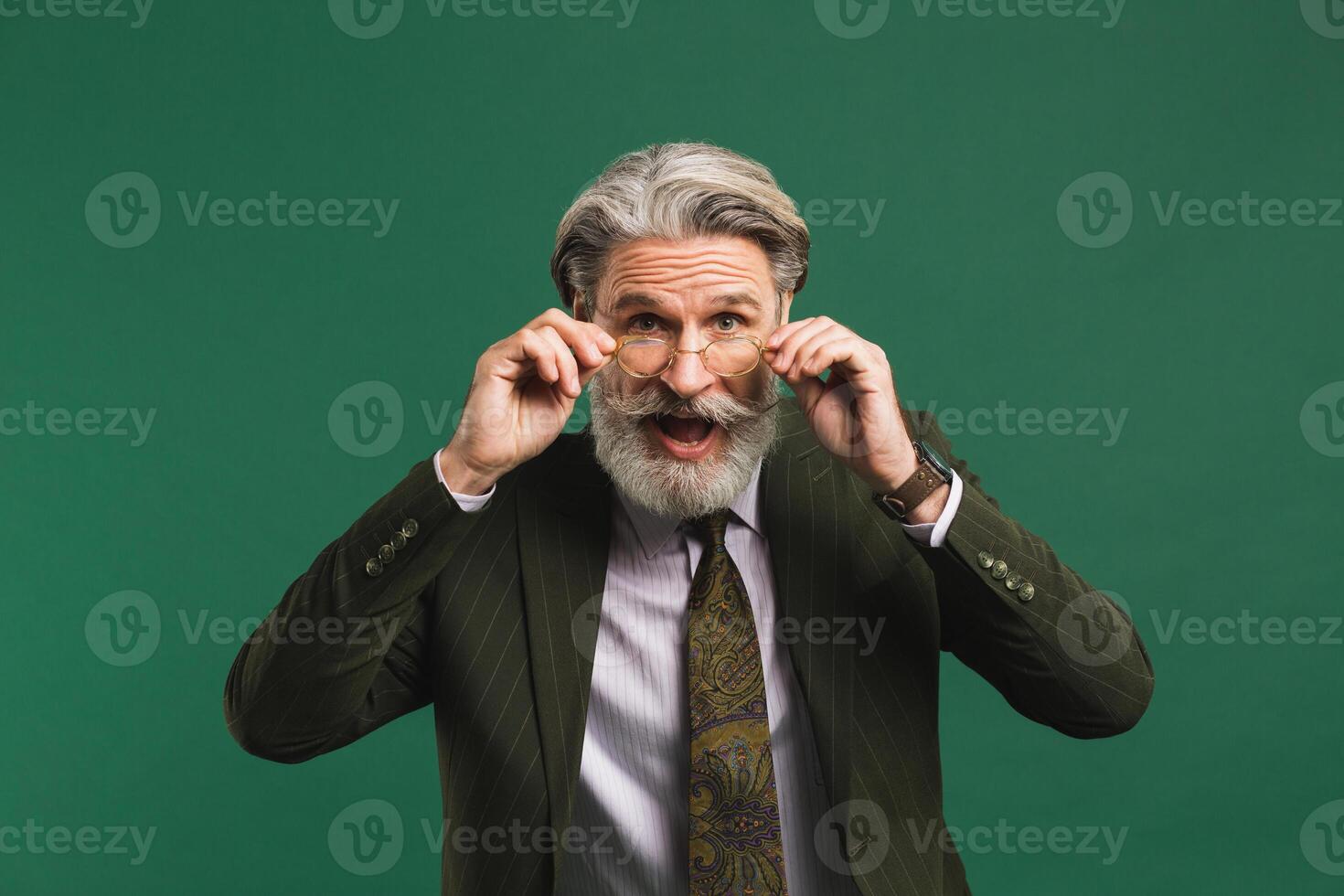 Bearded middle age teacher in khaki suit holds glasses with his hands and blinks his eyes with fun on green background photo