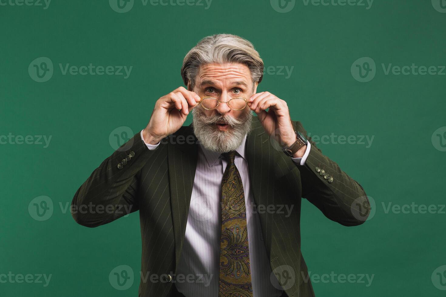 Bearded middle age teacher in khaki suit holds glasses with his hands and blinks his eyes with fun on green background photo