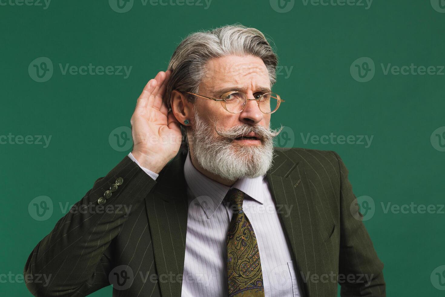Bearded middle aged teacher in khaki suit holds ear and eavesdrop on green background photo