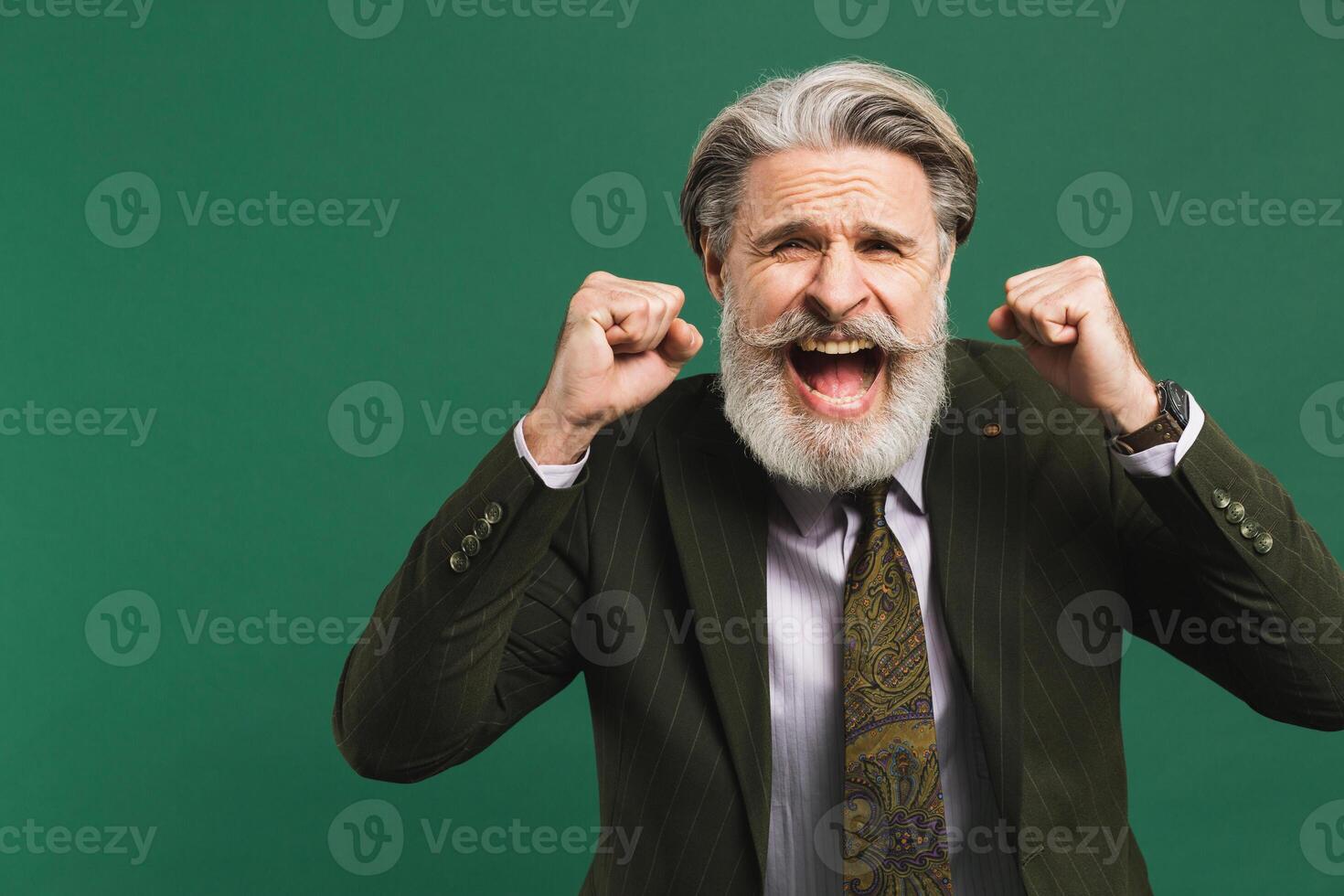 A middle-aged bearded man in a khaki suit shouts loudly and holds his fists against on a green background photo
