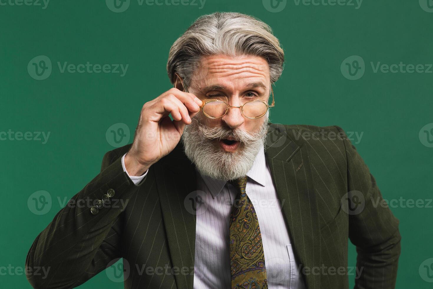 Bearded middle age teacher in khaki suit holds glasses with his hands and blinks his eyes with fun on green background photo
