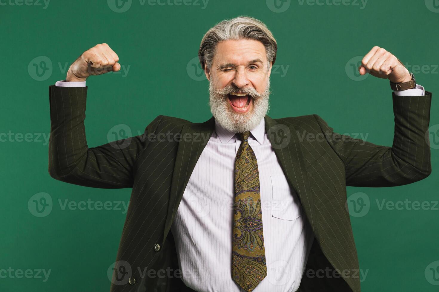 Bearded middle-aged man in khaki suit shows biceps and smiles emotionally on green background photo