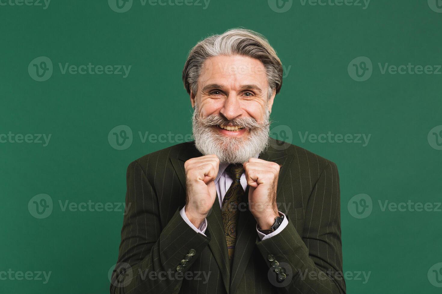 A middle-aged bearded man in a suit holds his fists, aches and rejoices on a green background. photo