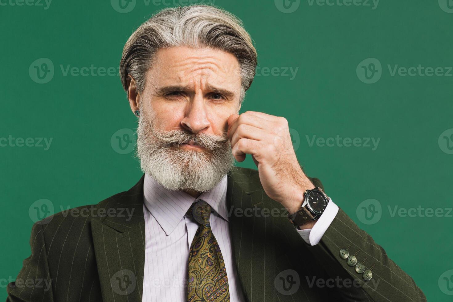 Stylish middle-aged man in khaki suit fixes mustache and beard on green background photo