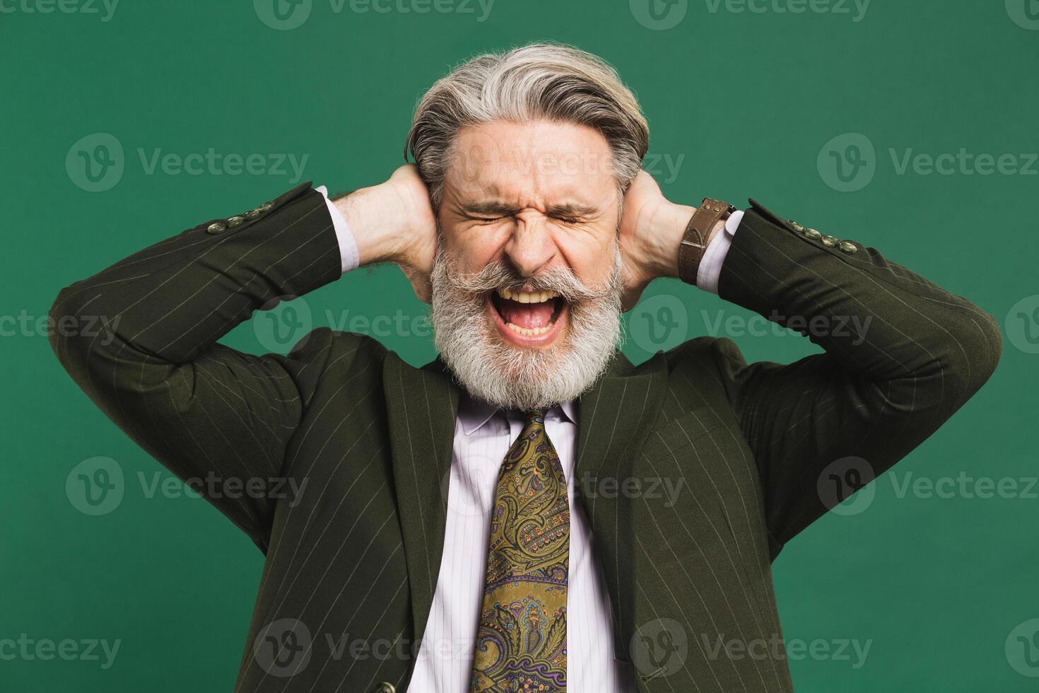 A bearded middle-aged teacher in a suit has stress and closes his ears with his hands and shouts with anger at the green background. photo
