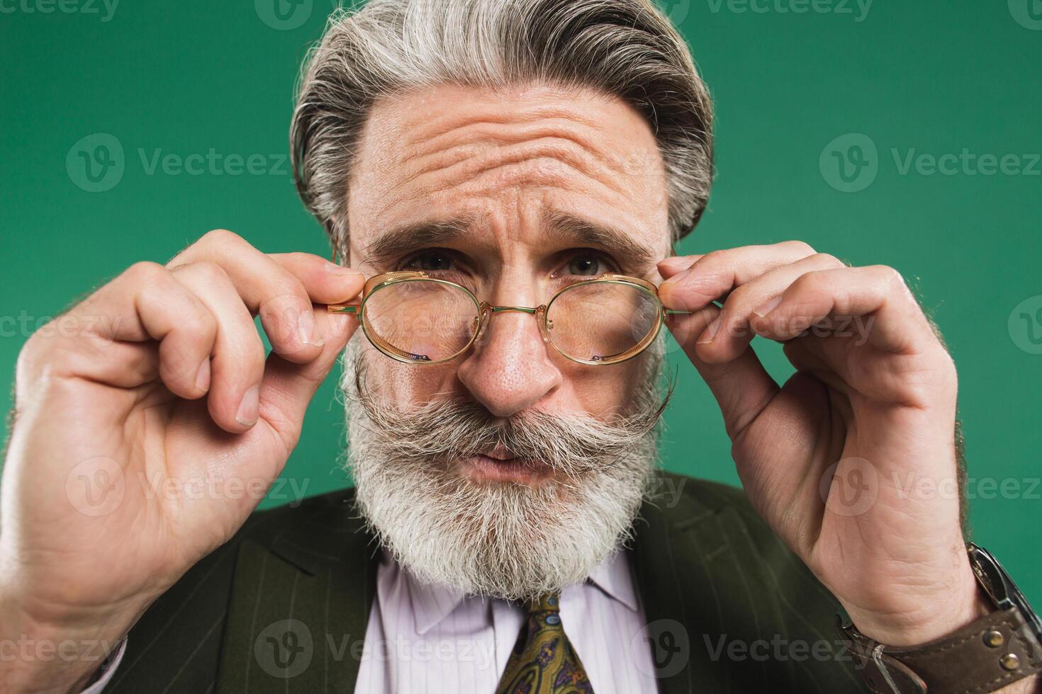 Bearded middle age teacher in khaki suit holds glasses with his hands and blinks his eyes with fun on green background photo
