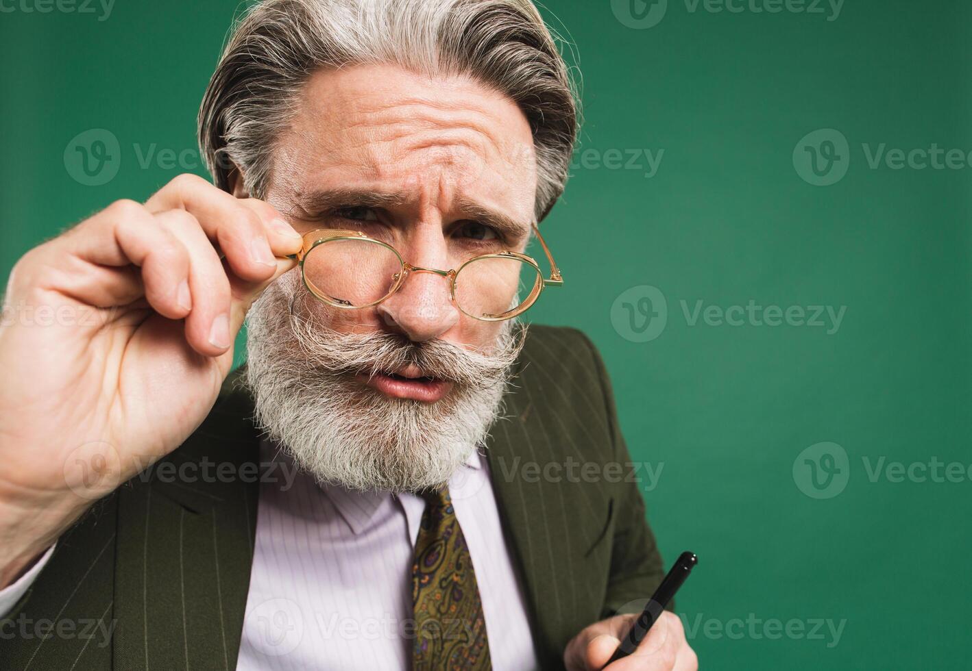 Bearded and mustachioed middle-aged teacher in khaki suit holds hand with glasses and looks closeup on green background photo