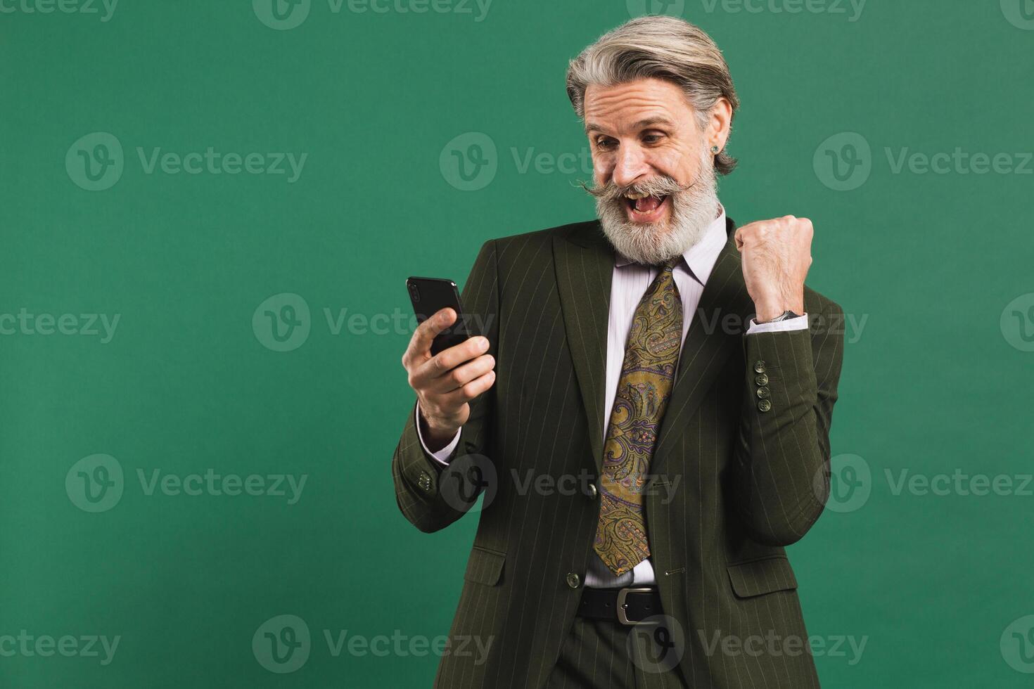Bearded middle-aged man in khaki suit screaming loudly and looking at phone while holding fist of happiness against green background photo