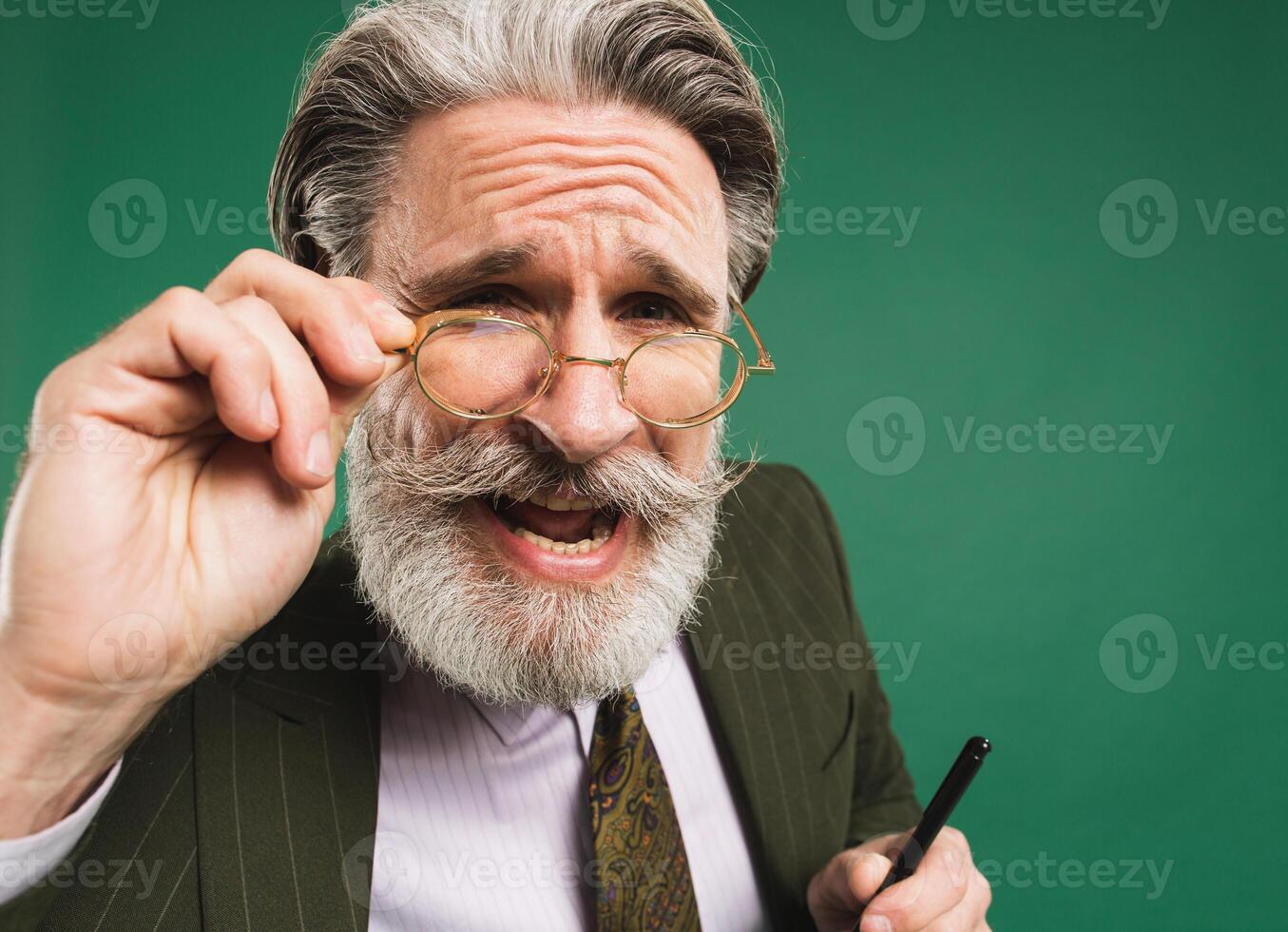 Bearded and mustachioed middle-aged teacher in khaki suit holds hand with glasses and looks close up and says something on green background photo