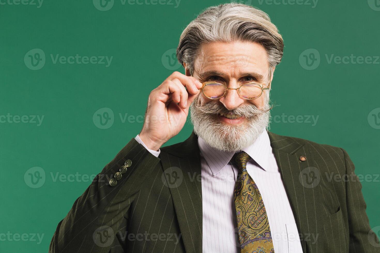 Stylish middle-aged bearded man in khaki suit holding glasses over green background photo