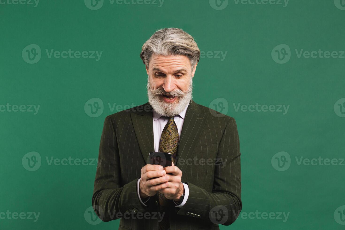 Bearded middle-aged man in khaki suit looks at phone and shows shock on green background photo