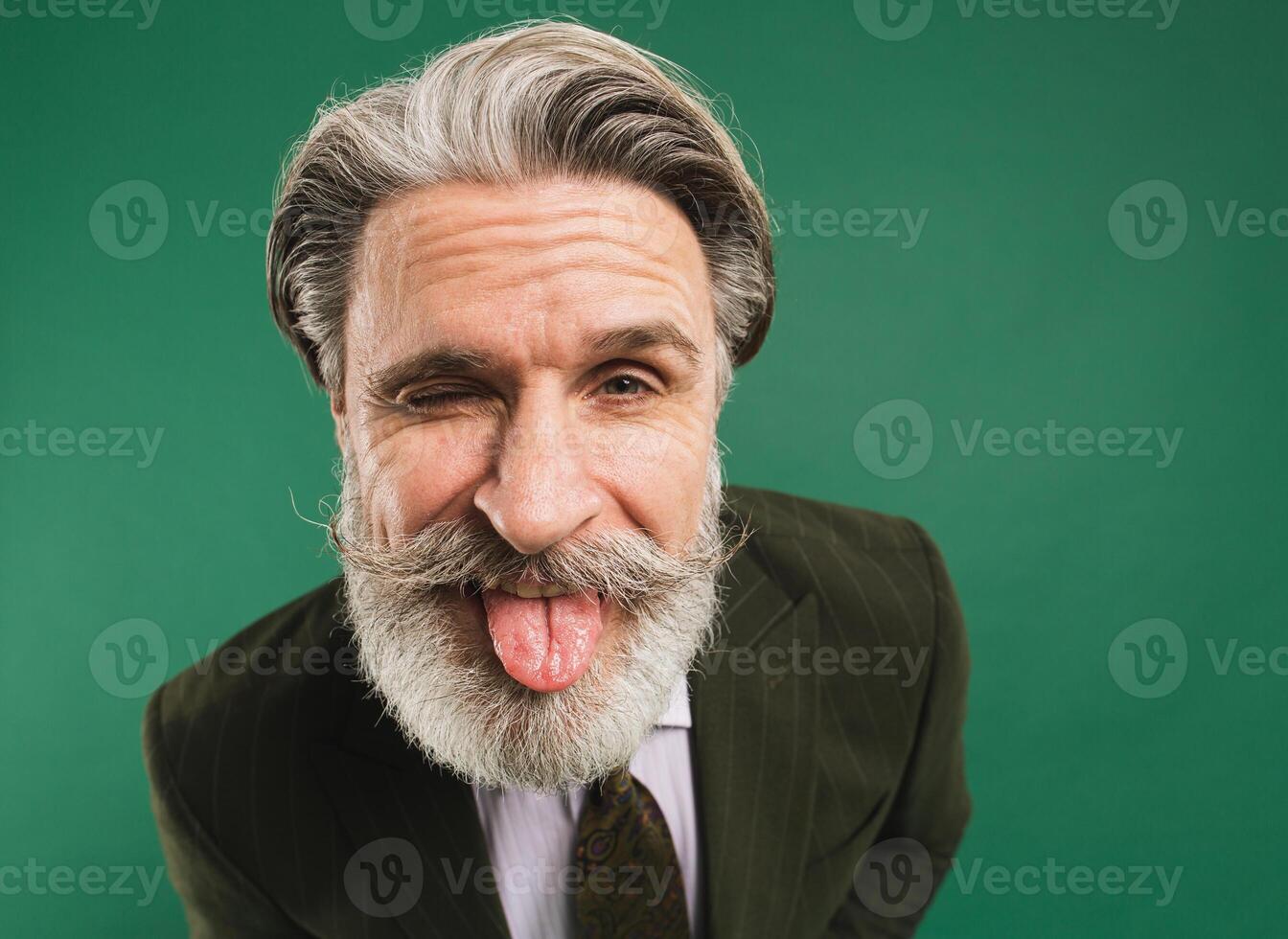 Stylish middle-aged bearded man in khaki suit shows tongue closeup on green background photo