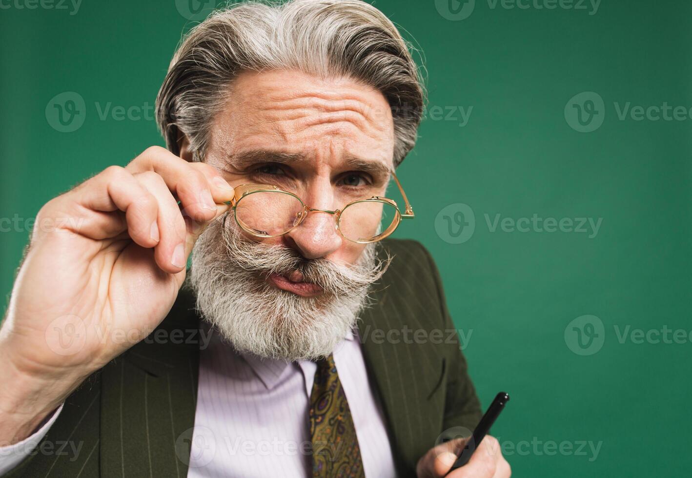 Bearded and mustachioed middle-aged teacher in khaki suit holds hand with glasses and looks closeup on green background photo