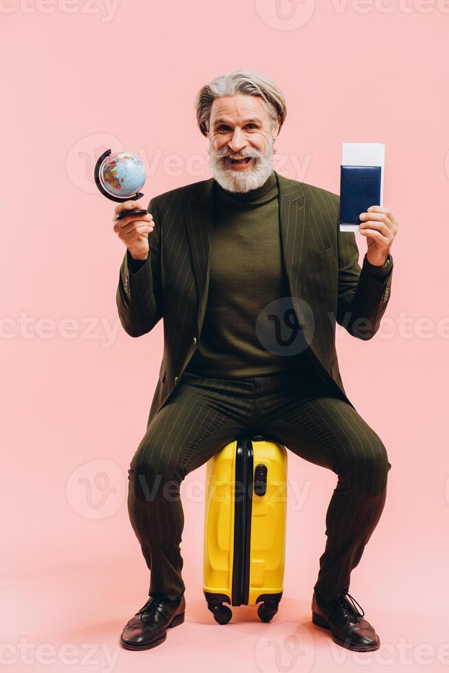 Stylish middle-aged man in a suit sits on a yellow suitcase and holds a passport and a globe. photo