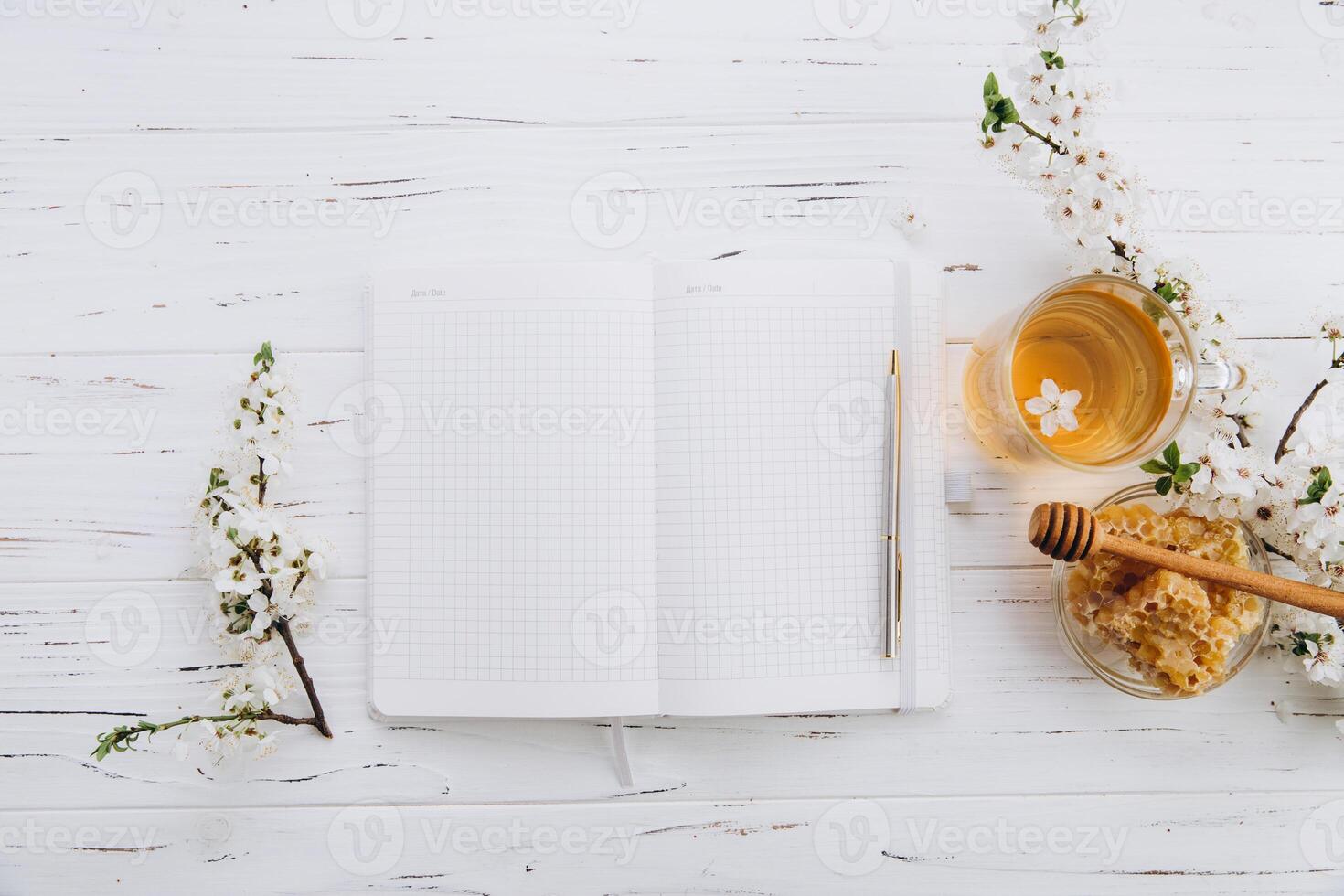 Spring composition. A cup of herbal tea, a jar of honey and cherry blossom near notebook on a white wooden background, top view. Place for text. Healthy food planning concept. photo