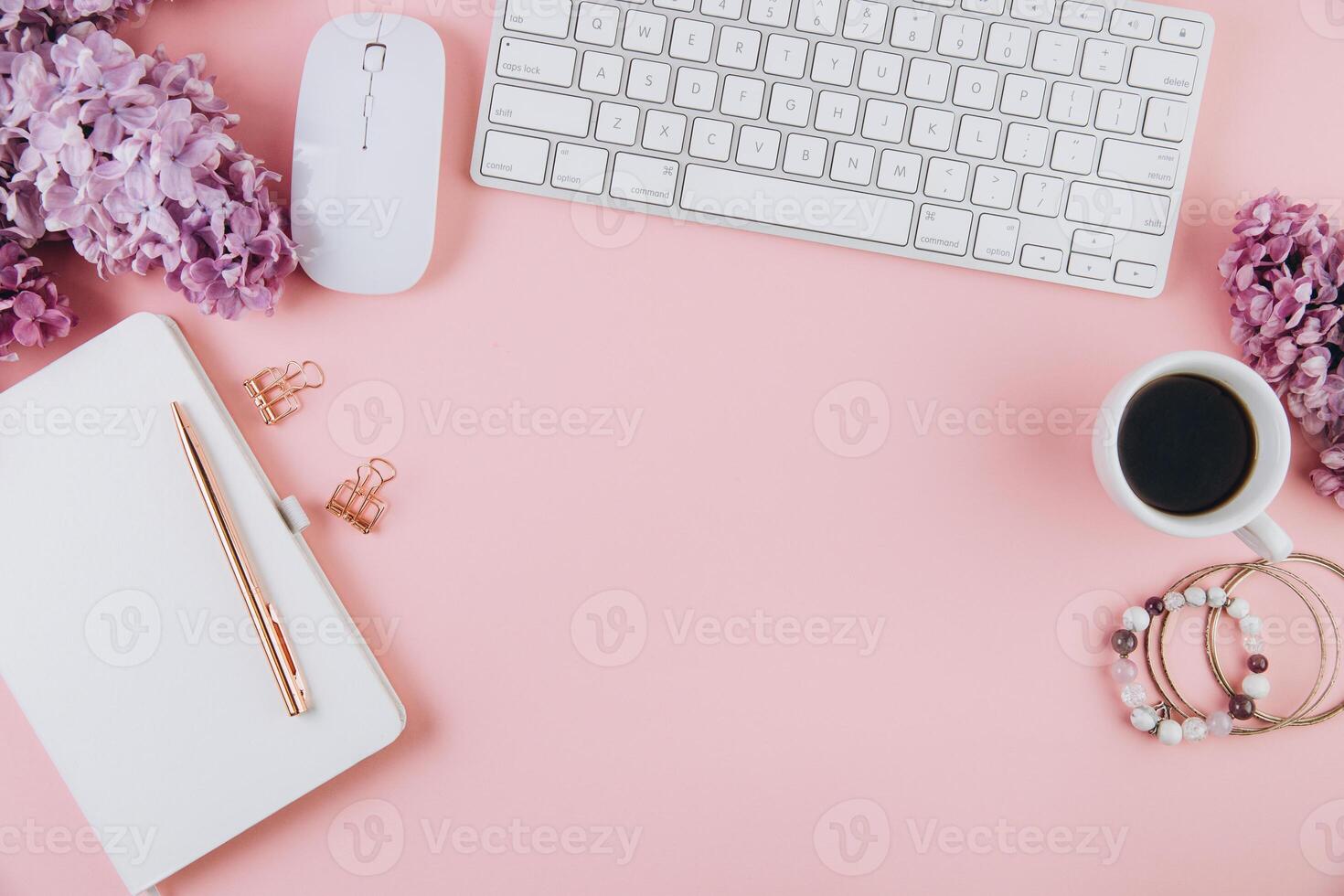 Spring flat lay top view home office workspace - modern keyboard, cup of coffee and notebook with lilac flowers on a pink desk background photo