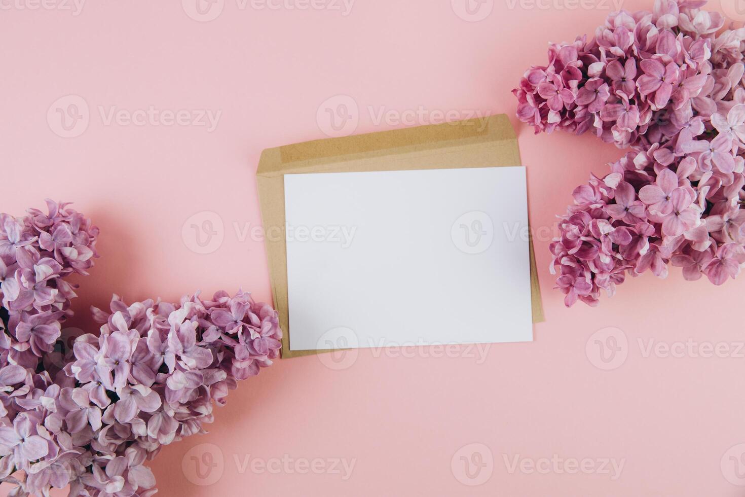 Flat lay of kraft envelope and blank paper card with copy space mock up Top view home desk workspace decorated with spring lilac flowers. photo