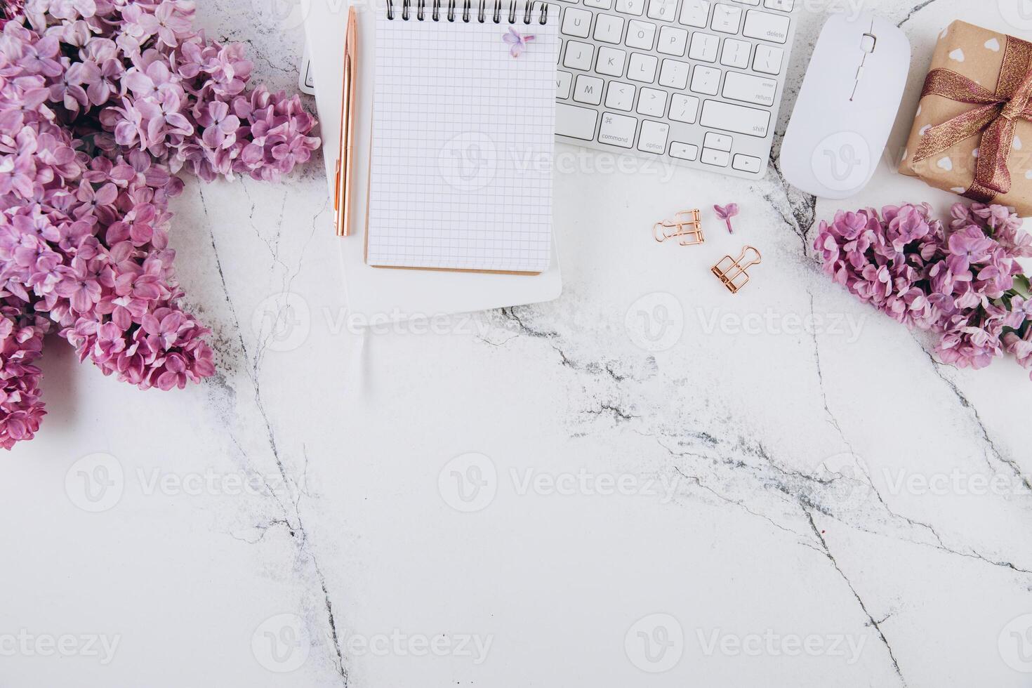 Spring flat lay top view home office workspace - modern keyboard, gift and notebook with lilac flowers on a marble desk background photo