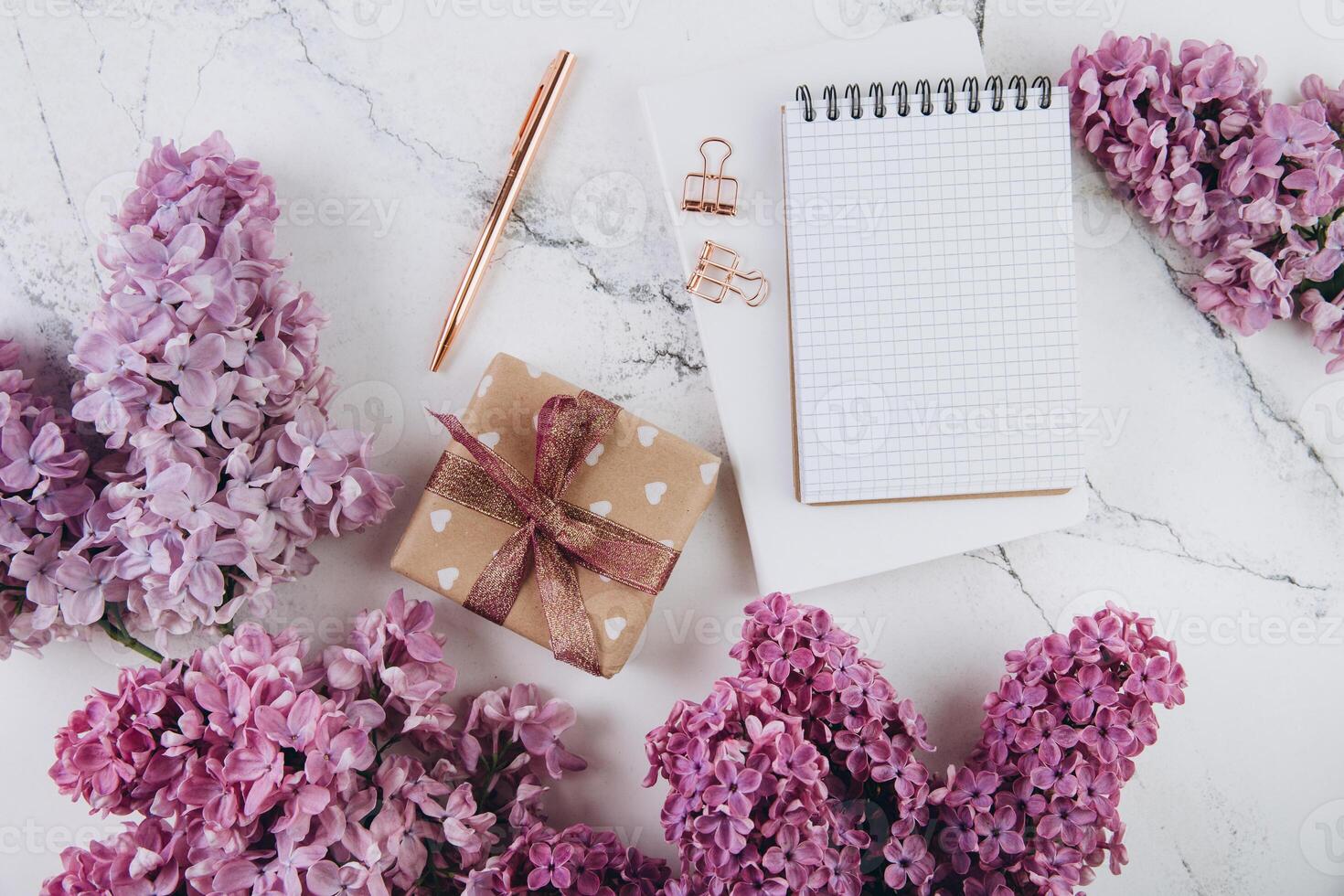 Spring flat lay top view home office workspace - gift and notebook with lilac flowers on a marble desk background photo