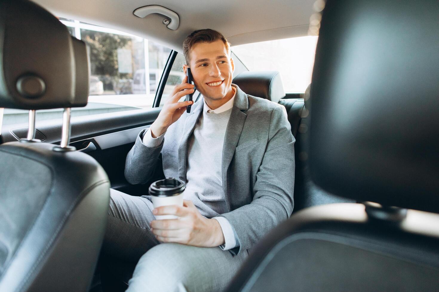 Young businessman talking on a mobile phone and drinking coffee while sitting in the back seat of a car. photo