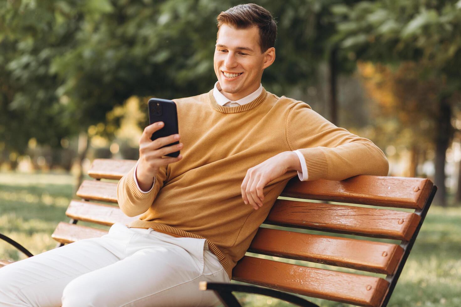 Modern handsome smiling man in yellow and white clothes talking on the phone sitting on a park bench at sunset photo