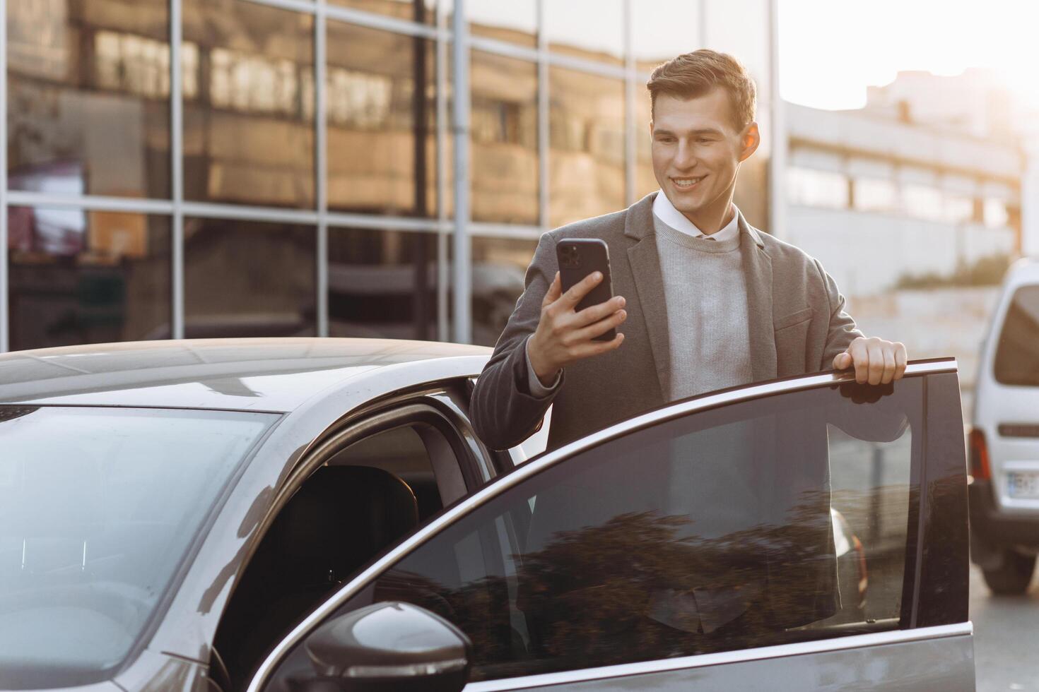 Handsome modern young man with phone entering his car while standing outdoors photo