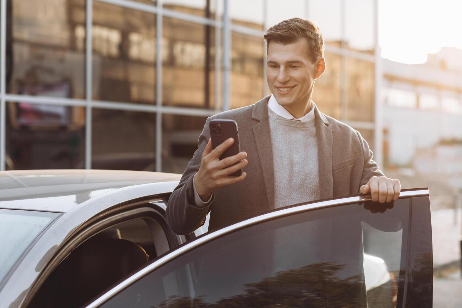 Handsome modern young man with phone entering his car while standing outdoors photo