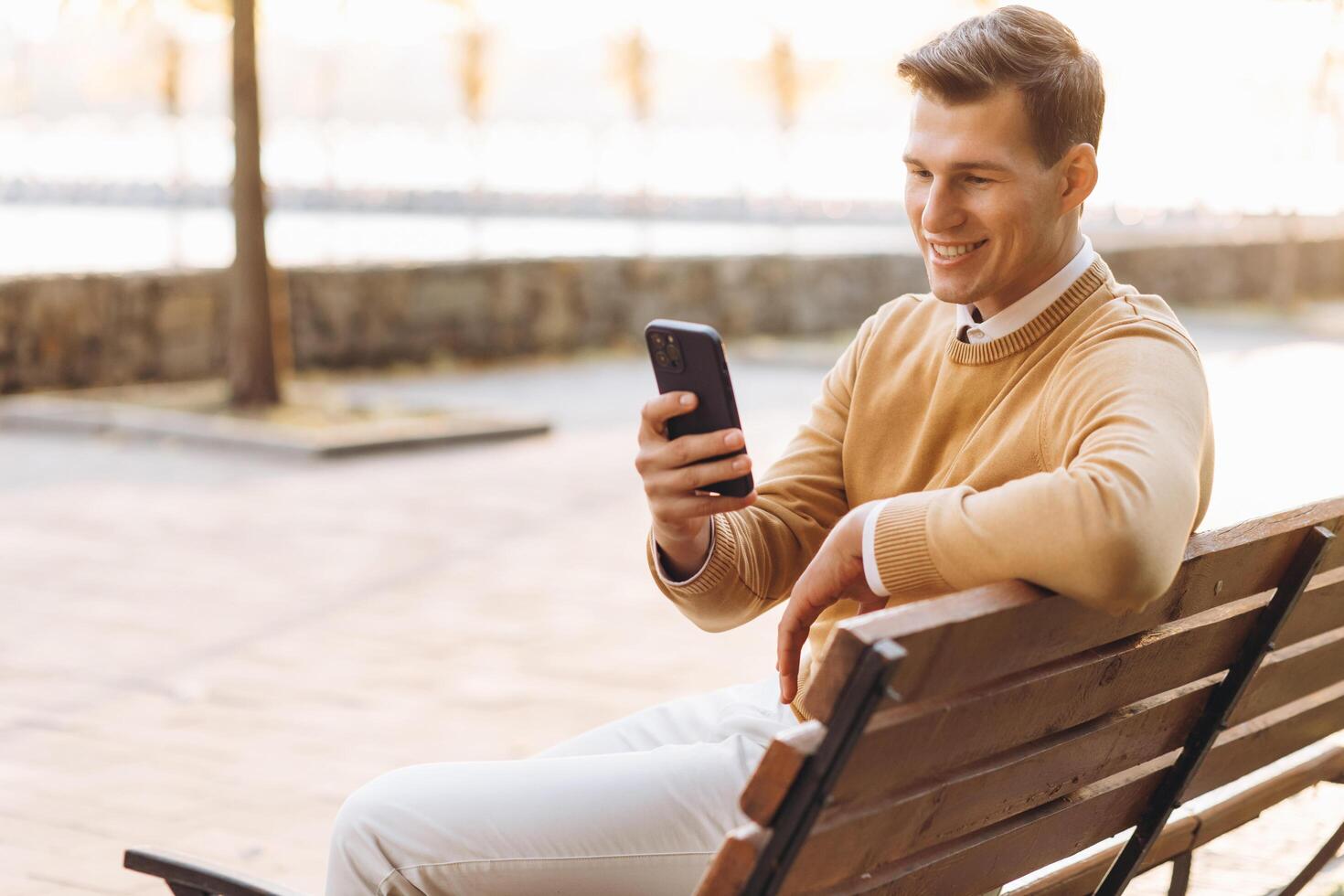 Modern handsome smiling man in yellow and white clothes talking on the phone sitting on a park bench at sunset photo