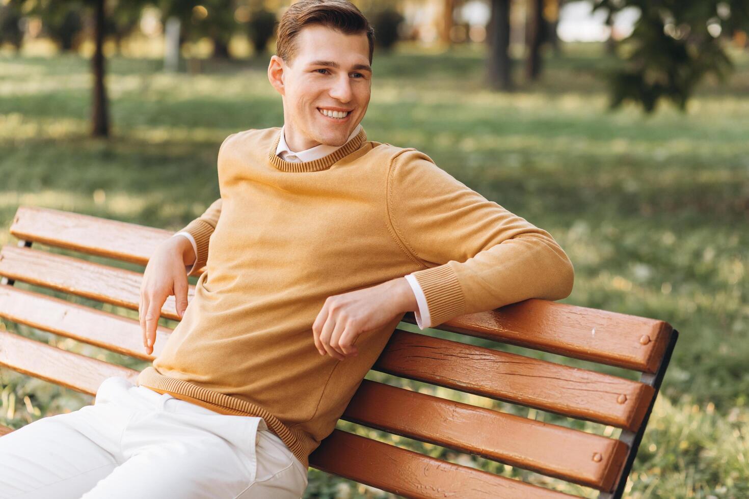 Modern handsome smiling man in yellow and white clothes sitting on a park bench at sunset photo