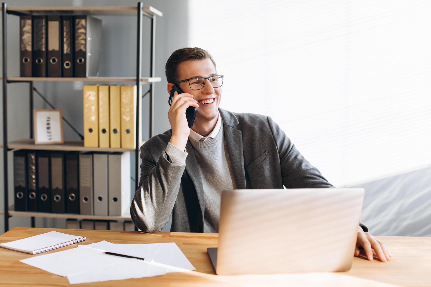Modern man with glasses smiling, office worker working on laptop and having a phone conversation photo
