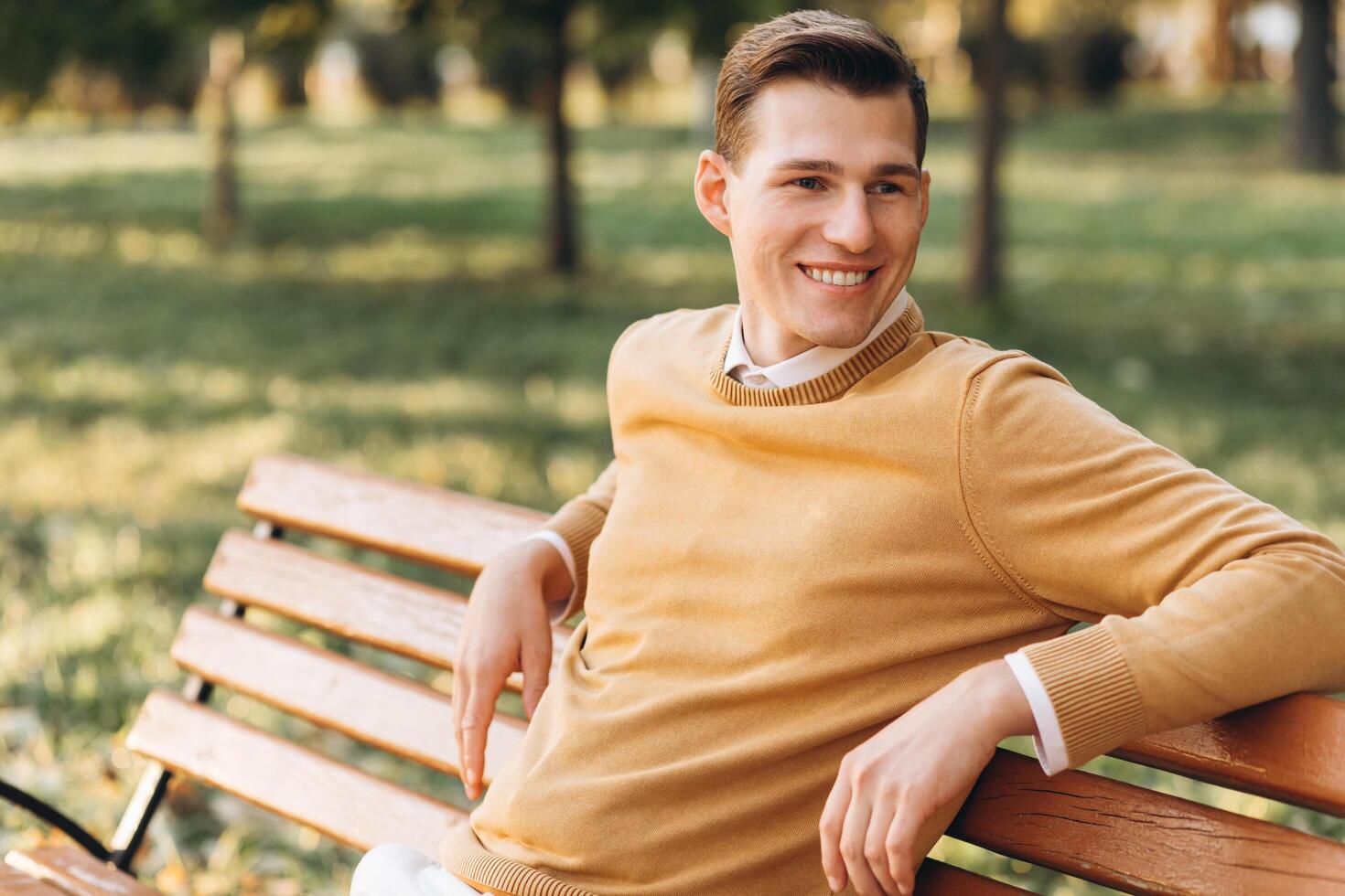 Modern handsome smiling man in yellow and white clothes sitting on a park bench at sunset photo