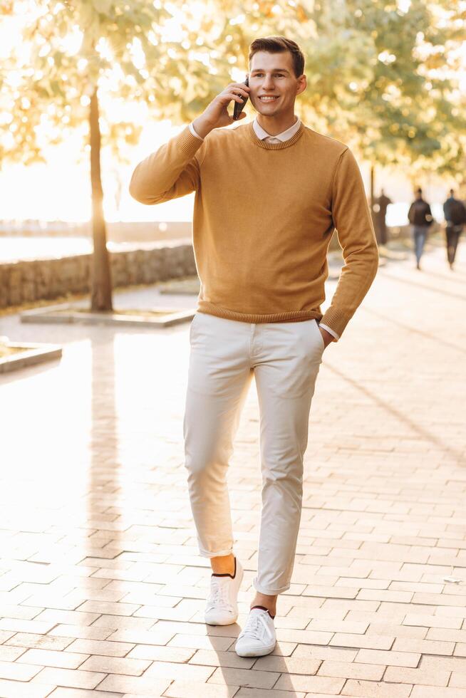 Modern handsome smiling man in yellow and white clothes talking on the phone in the park at sunset photo