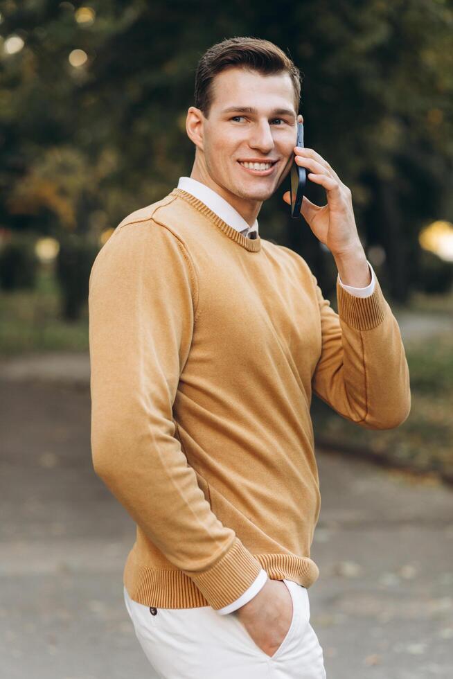 Modern handsome smiling man in yellow and white clothes talking on the phone in the park at sunset photo