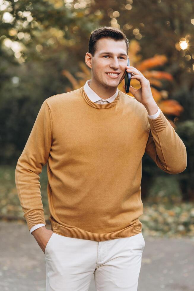 Modern handsome smiling man in yellow and white clothes talking on the phone in the park at sunset photo