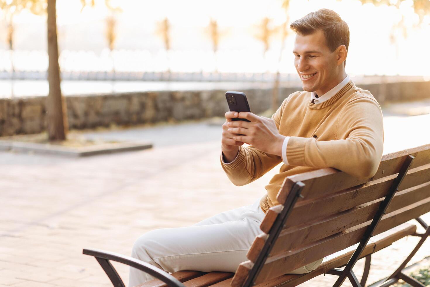 Modern handsome smiling man in yellow and white clothes talking on the phone sitting on a park bench at sunset photo