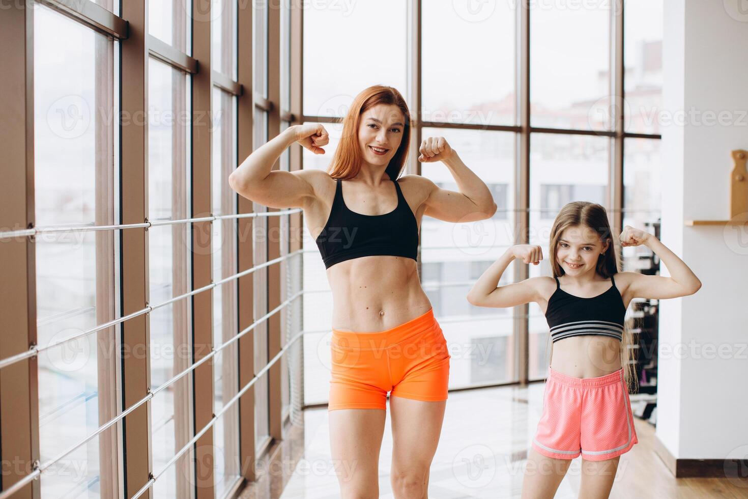 Beautiful strong woman and charming little daughter are showing their biceps and smiling while working out at gym photo