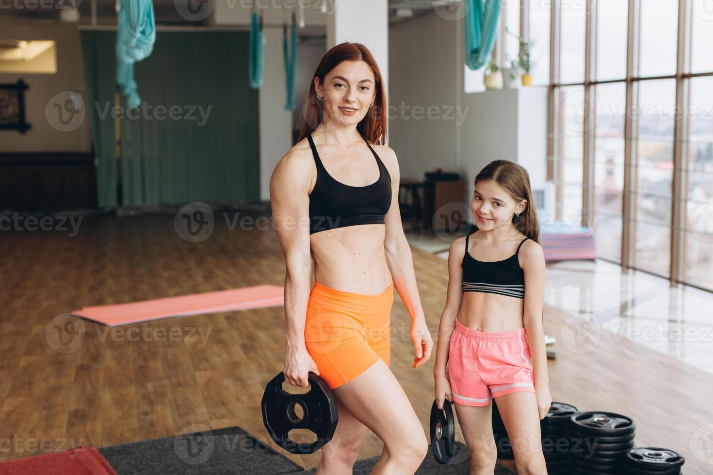 Mom and Daughter are doing exercises. Family in a gym. Little girl with strong mother are training with pancakes rods photo
